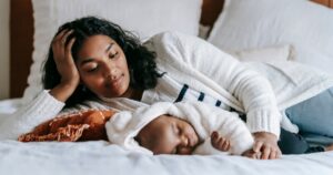 The Mother’s Day Paradox: The Challenge of Invisible Motherhood - Single Mama Way Counselling Mother and baby resting together on a bed, she gently supports the baby with a sweatered arm in a cozy, calm moment.