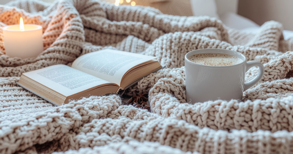 Winter Self-Care for Single Mums: How to Create a Domestic Sanctuary - Single Mama Way Counselling - Single Mama Way Counselling Cozy reading setup with an open book, a mug of coffee, and a lit candle on a chunky beige knit blanket.